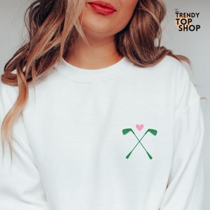 May include: White sweatshirt with a green golf club design and a pink heart.