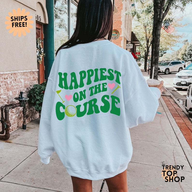 May include: White sweatshirt with green text that says "Happiest on the Course" with a golf ball and golf club graphics. The text is in a retro font style with pink hearts.