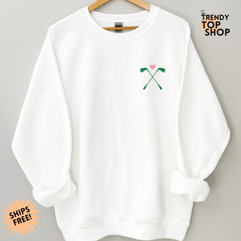 May include: White sweatshirt with a green golf club design and a pink heart on the front. The text "The Trendy Top Shop" is in the top right corner of the image.