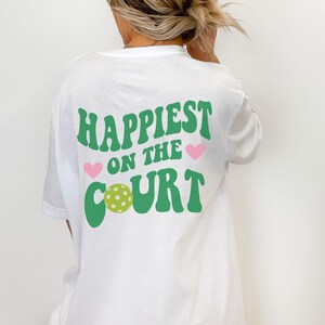 May include: White t-shirt with green text that says "Happiest on the Court" with a green pickleball and pink hearts.
