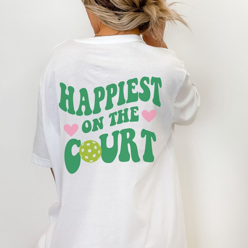 May include: White t-shirt with green text that says "Happiest on the Court" with a green pickleball and pink hearts.