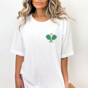 May include: White t-shirt with a green pickleball paddle and pink ball graphic.