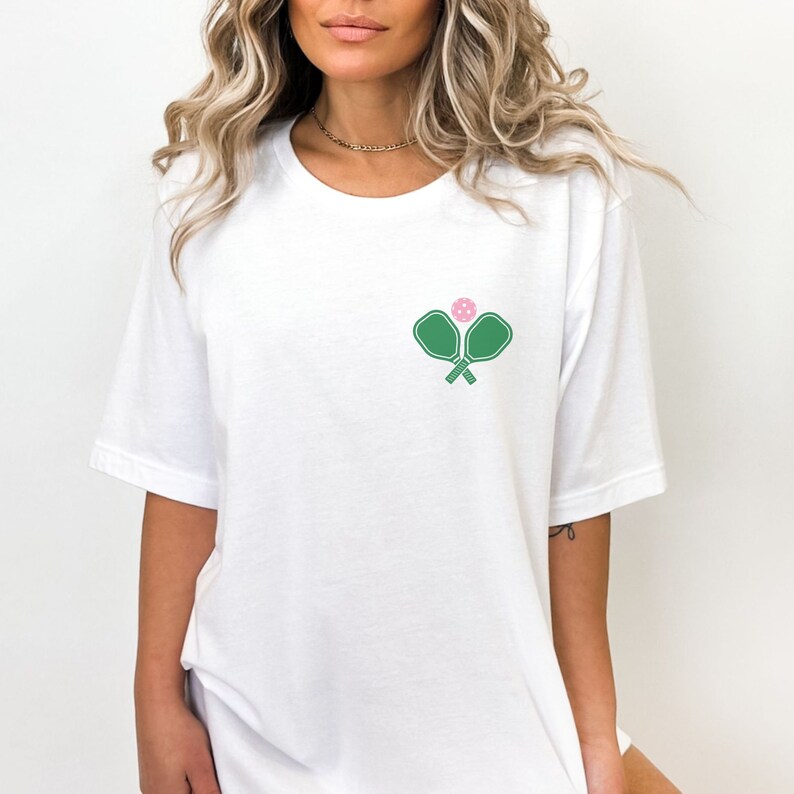 May include: White t-shirt with a green pickleball paddle and pink ball graphic.