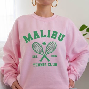 K&ouml;nnte beinhalten: Rosafarbener Pullover mit einem gr&uuml;nen Aufdruck, der "Malibu Tennis Club" mit einem Tennisschl&auml;ger und einem Ball zeigt.