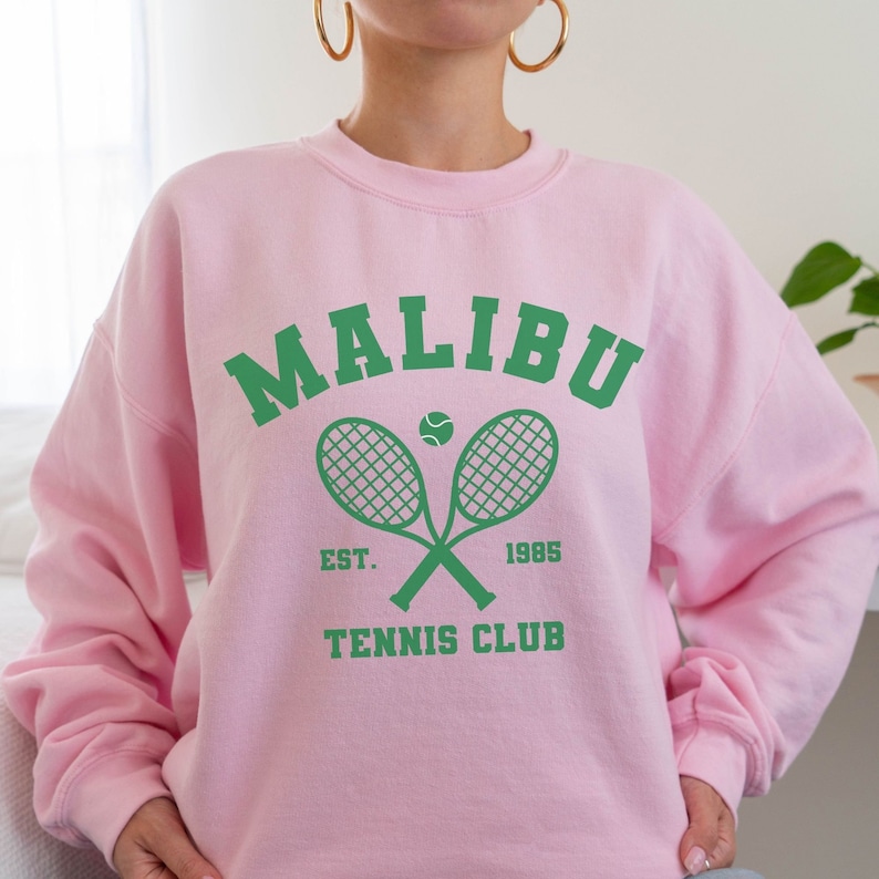 K&ouml;nnte beinhalten: Rosafarbener Pullover mit einem gr&uuml;nen Aufdruck, der "Malibu Tennis Club" mit einem Tennisschl&auml;ger und einem Ball zeigt.