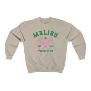 K&ouml;nnte beinhalten: Ein beiger Pullover mit einem gr&uuml;nen und rosafarbenen Tennisclub-Logo. Das Logo lautet "Malibu Tennis Club Est. 1965" mit zwei pinkfarbenen Tennisschl&auml;gern, die &uuml;ber einem Tennisball gekreuzt sind.
