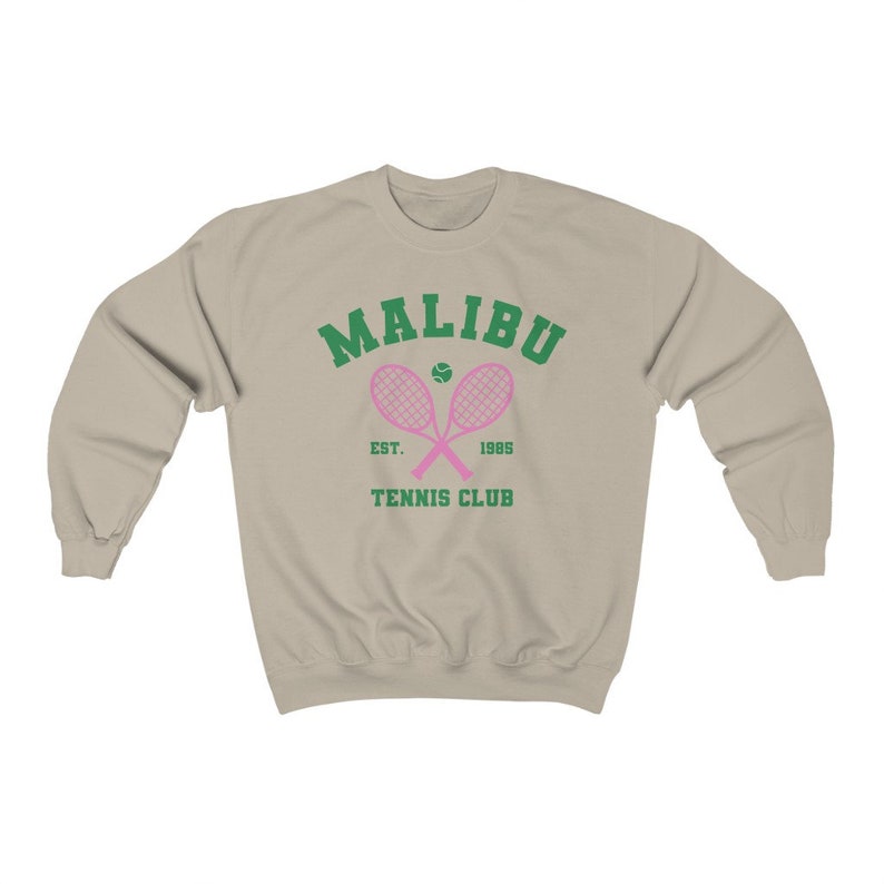 K&ouml;nnte beinhalten: Ein beiger Pullover mit einem gr&uuml;nen und rosafarbenen Tennisclub-Logo. Das Logo lautet "Malibu Tennis Club Est. 1965" mit zwei pinkfarbenen Tennisschl&auml;gern, die &uuml;ber einem Tennisball gekreuzt sind.