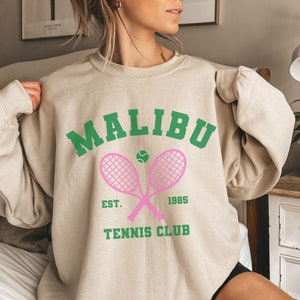 K&ouml;nnte beinhalten: Ein beiger Pullover mit einem gr&uuml;n-rosa Grafikdesign. Die Grafik lautet "Malibu Tennis Club" mit dem Text "Est. 1985" darunter. Die Grafik zeigt auch zwei rosa Tennisschl&auml;ger, die sich &uuml;berkreuzen, mit einem gr&uuml;nen Tennisball in der Mitte.