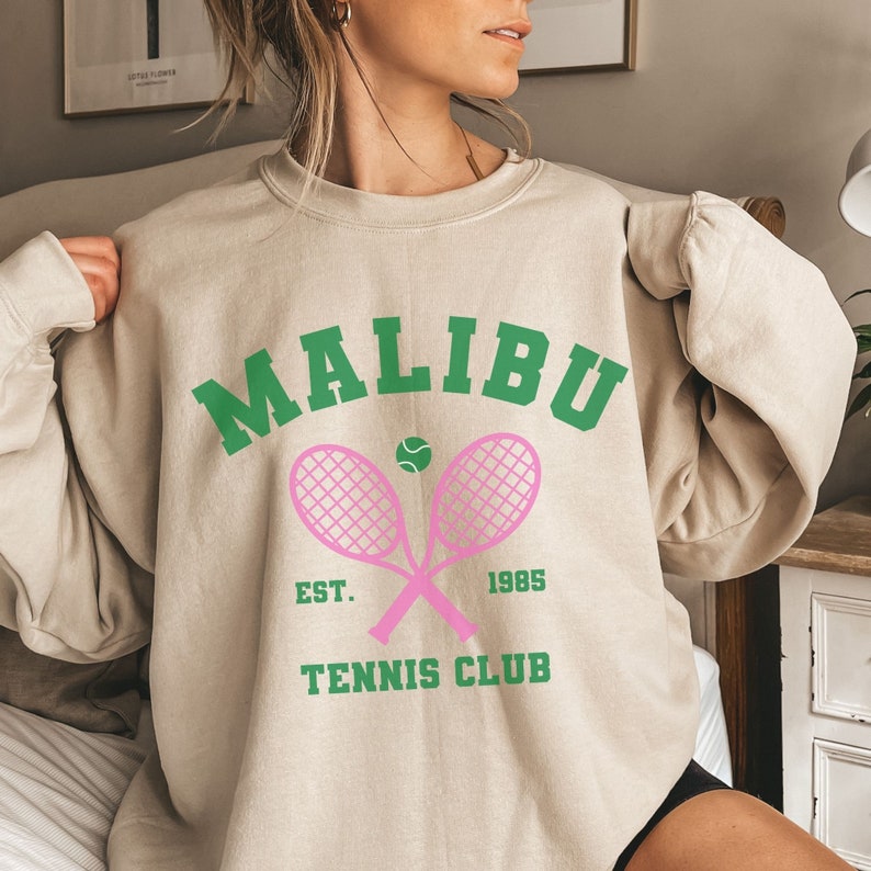 K&ouml;nnte beinhalten: Ein beiger Pullover mit einem gr&uuml;n-rosa Grafikdesign. Die Grafik lautet "Malibu Tennis Club" mit dem Text "Est. 1985" darunter. Die Grafik zeigt auch zwei rosa Tennisschl&auml;ger, die sich &uuml;berkreuzen, mit einem gr&uuml;nen Tennisball in der Mitte.