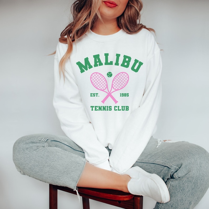 K&ouml;nnte beinhalten: Wei&szlig;er Pullover mit einem gr&uuml;nen und rosafarbenen Grafikdesign, auf dem "Malibu Tennis Club Est. 1985" mit zwei rosafarbenen Tennisschl&auml;gern und einem gr&uuml;nen Tennisball steht.