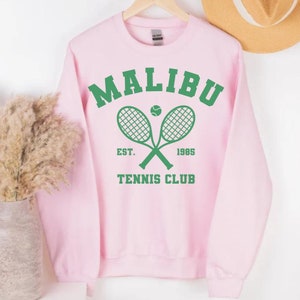 K&ouml;nnte beinhalten: Rosafarbener Pullover mit einer gr&uuml;nen Grafik von zwei gekreuzten Tennisschl&auml;gern mit einem Tennisball in der Mitte. Die Grafik lautet "Malibu Tennis Club Est. 1985"