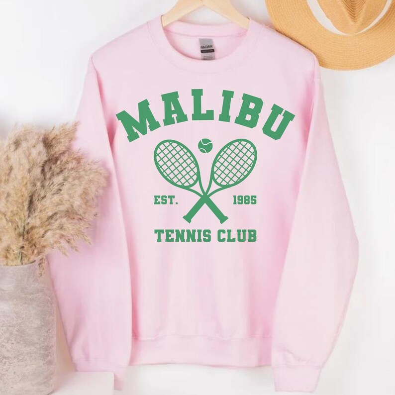K&ouml;nnte beinhalten: Rosafarbener Pullover mit einer gr&uuml;nen Grafik von zwei gekreuzten Tennisschl&auml;gern mit einem Tennisball in der Mitte. Die Grafik lautet "Malibu Tennis Club Est. 1985"