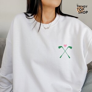May include: White sweatshirt with a green golf club design and a pink heart. The text "The Trendy Top Shop" is in the upper right corner of the image.