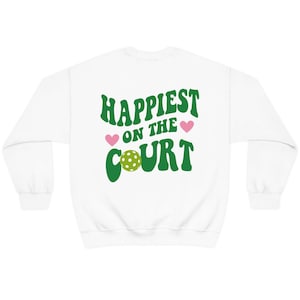 May include: White sweatshirt with green text that reads "Happiest on the Court" with a green pickleball and pink hearts.