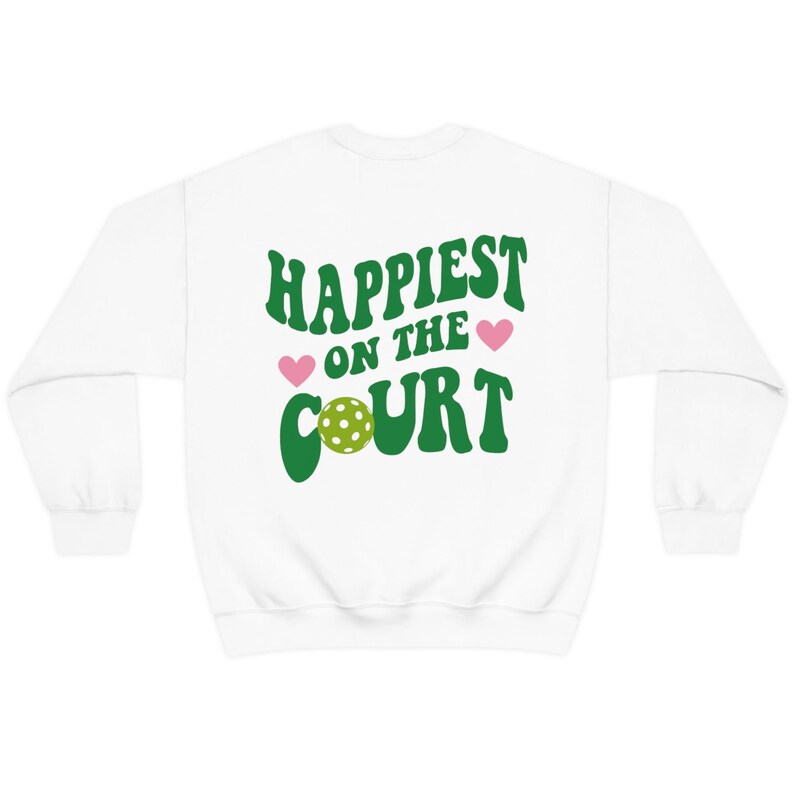 May include: White sweatshirt with green text that reads "Happiest on the Court" with a green pickleball and pink hearts.