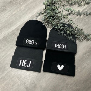 Könnte beinhalten: Vier schwarze Strickmützen mit weiß gesticktem Text. Die Mützen zeigen die Wörter "ciao kakao", "moin", "hej" und ein weißes Herz.