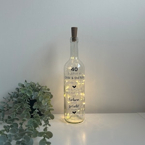 Könnte beinhalten: Eine durchsichtige Glasflasche mit einem Korkverschluss und weißen Lichtern im Inneren. Die Flasche hat ein schwarzes Etikett mit dem Text "40 Jahre Gabi & Dieter 05.07.1984 - 05.07.2025" und "Das schönste hier auf Erden ist lieben und geliebt zu werden. Alles Gute zur Silberhochzeit."