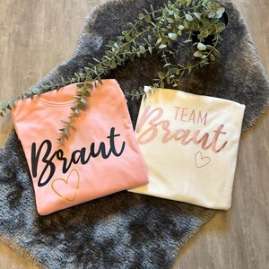 May include: Two pink and white t-shirts with the text "Braut" and "Team Braut" in black and gold lettering. The shirts are on a gray fuzzy surface.