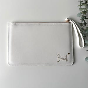 Könnte beinhalten: Eine weiße, rechteckige Handgelenktasche mit strukturierter Oberfläche und goldenem Reißverschluss. Das Wort Braut ist in Schreibschrift in der unteren rechten Ecke aufgedruckt, ergänzt durch ein kleines goldenes Herzsymbol über dem Buchstaben t.