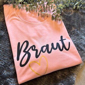 May include: A pink t-shirt with the word "Braut" in black lettering and a gold heart.