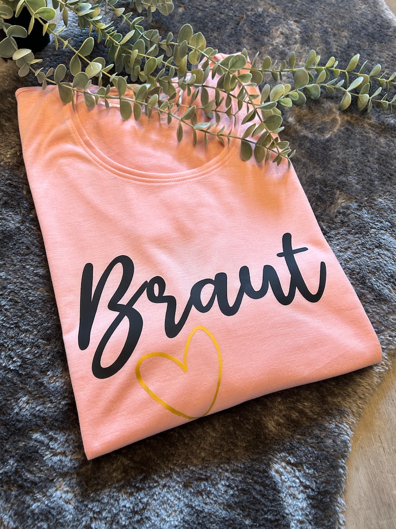 May include: A pink t-shirt with the word "Braut" in black lettering and a gold heart.