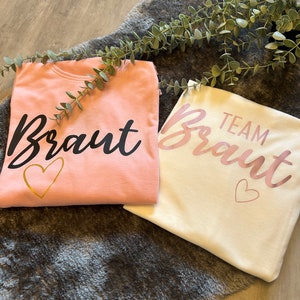 May include: Two t-shirts, one pink and one white, with the word "Braut" written on them. The pink shirt has a gold heart and the white shirt has a pink heart. The text "Team" is written above the word "Braut" on the white shirt.