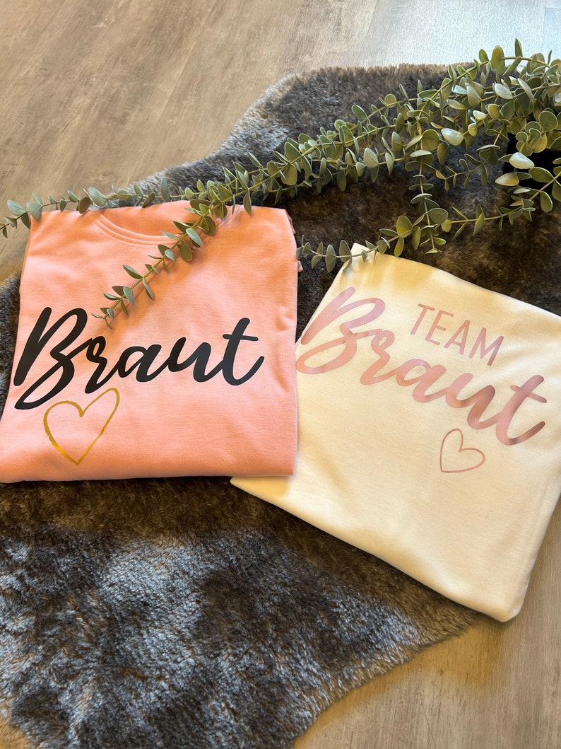 May include: Two t-shirts, one pink and one white, with the word "Braut" written on them. The pink shirt has a gold heart and the white shirt has a pink heart. The text "Team" is written above the word "Braut" on the white shirt.