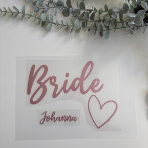 以下が含まれることがあります： 「Bride」という単語が筆記体で書かれたピンクのビニールステッカー、ハート、そしてその下に「Johanna」という名前が書かれています。