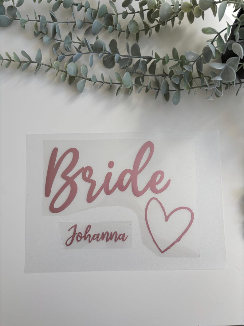 以下が含まれることがあります： 「Bride」という単語が筆記体で書かれたピンクのビニールステッカー、ハート、そしてその下に「Johanna」という名前が書かれています。