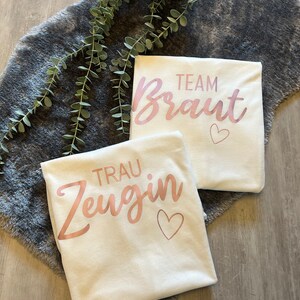 May include: Two white t-shirts with rose gold lettering. One shirt says "Team Braut" with a heart and the other says "Trau Zeugin" with a heart.