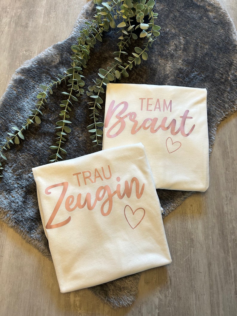 May include: Two white t-shirts with rose gold lettering. One shirt says "Team Braut" with a heart and the other says "Trau Zeugin" with a heart.