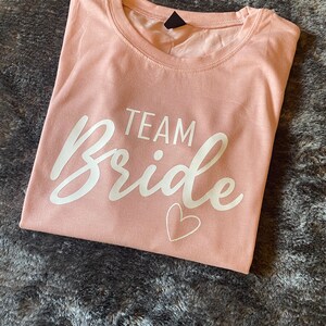 以下が含まれることがあります： 薄いピンクのTシャツに、白い文字で「TEAM Bride」と小さな白いハートが書かれています。