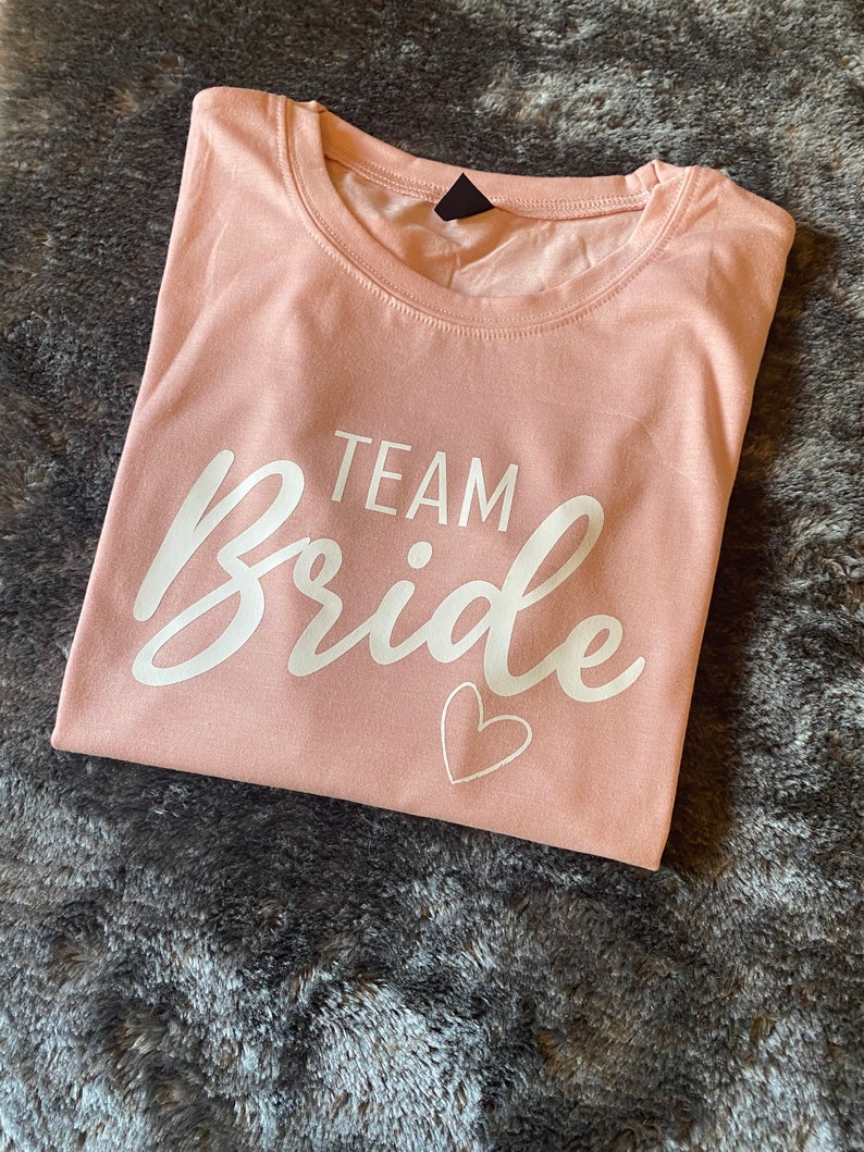 以下が含まれることがあります： 薄いピンクのTシャツに、白い文字で「TEAM Bride」と小さな白いハートが書かれています。