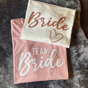 以下が含まれることがあります： 白いTシャツとピンクのTシャツ2枚で、それぞれに「Bride」という単語が筆記体で書かれています。白いTシャツには、「Bride」という単語がローズゴールドの文字で書かれており、その下にハートがあります。ピンクのTシャツには、「Bride」という単語が白い文字で書かれており、その上に「Team」という単語があり、その下にハートがあります。