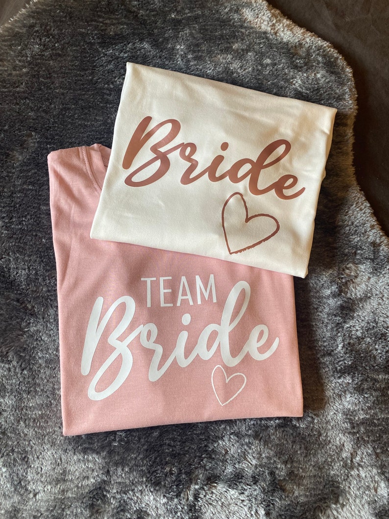 以下が含まれることがあります： 白いTシャツとピンクのTシャツ2枚で、それぞれに「Bride」という単語が筆記体で書かれています。白いTシャツには、「Bride」という単語がローズゴールドの文字で書かれており、その下にハートがあります。ピンクのTシャツには、「Bride」という単語が白い文字で書かれており、その上に「Team」という単語があり、その下にハートがあります。