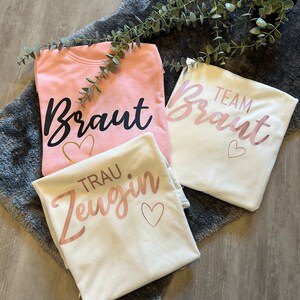 May include: Three white t-shirts with pink and black lettering. One shirt says "Braut" with a gold heart, another says "Team Braut" with a pink heart, and the last says "Trau Zeugin" with a pink heart.