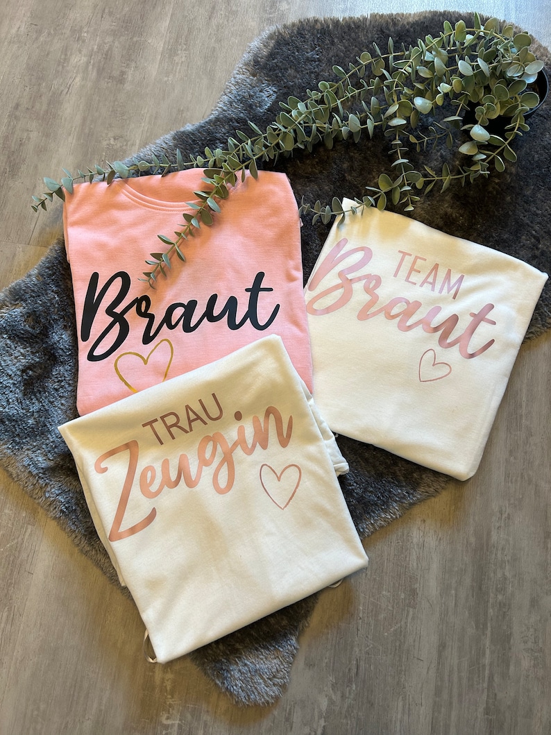 May include: Three white t-shirts with pink and black lettering. One shirt says "Braut" with a gold heart, another says "Team Braut" with a pink heart, and the last says "Trau Zeugin" with a pink heart.
