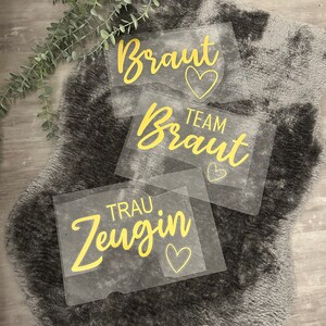 May include: Three clear plastic signs with gold lettering and hearts. The signs read "Braut", "Team Braut", and "Trau Zeugin".