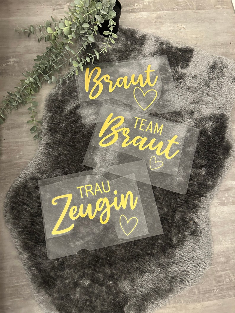 May include: Three clear plastic signs with gold lettering and hearts. The signs read "Braut", "Team Braut", and "Trau Zeugin".