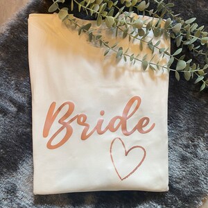 以下が含まれることがあります： 白いTシャツに、ローズゴールドの文字で「Bride」と、ローズゴールドのハートが書かれています。