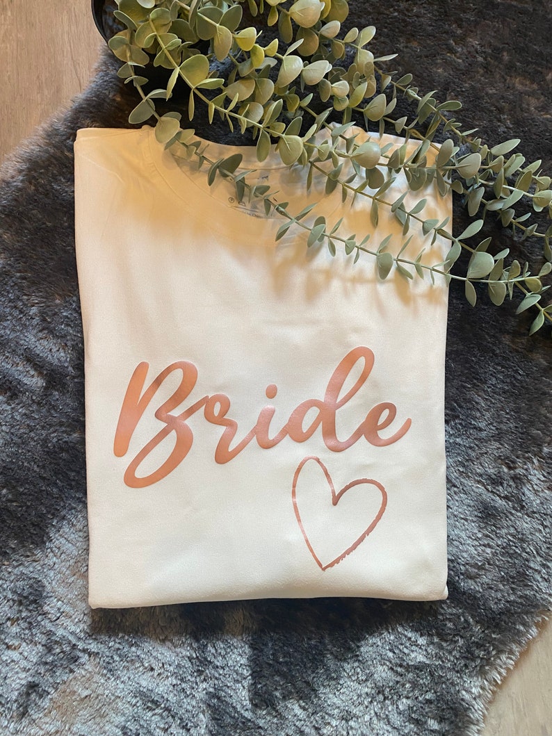 以下が含まれることがあります： 白いTシャツに、ローズゴールドの文字で「Bride」と、ローズゴールドのハートが書かれています。