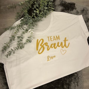 May include: White t-shirt with gold lettering that says "Team Braut Lisa" with a small heart.