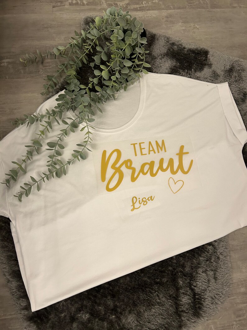May include: White t-shirt with gold lettering that says "Team Braut Lisa" with a small heart.