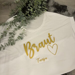 May include: White t-shirt with gold lettering that says "Braut" with a heart and the name "Tanja" underneath.