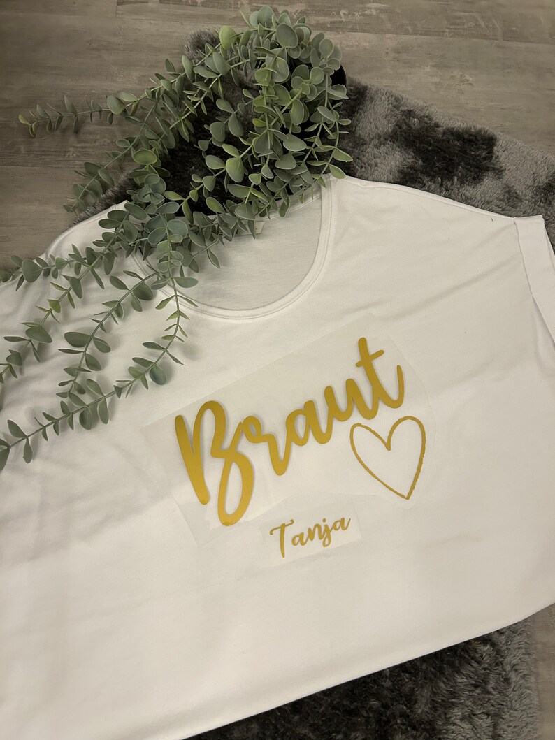 May include: White t-shirt with gold lettering that says "Braut" with a heart and the name "Tanja" underneath.