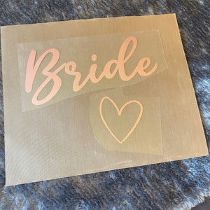 以下が含まれることがあります： 透明な転写紙のシートに、ローズゴールドのスクリプトで「Bride」という言葉と、ローズゴールドのハートが書かれています。