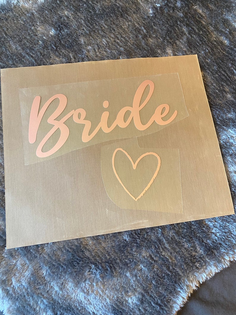 以下が含まれることがあります： 透明な転写紙のシートに、ローズゴールドのスクリプトで「Bride」という言葉と、ローズゴールドのハートが書かれています。