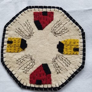May include: An octagonal, cream-coloured felt coaster with a black stitched border. It features red and yellow felt house appliqués with black accents, and embroidered wheat stalks. The coaster is handmade.