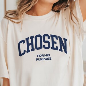 May include: A white t-shirt with a navy blue arch design that says "CHOSEN" and "FOR HIS PURPOSE" in smaller text below.