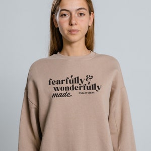 Pu&ograve; includere: Una felpa color marrone chiaro con il testo "fearfully & wonderfully made" in lettere nere. Il testo &egrave; in uno stile di font retr&ograve;. Il testo "PSALM 139:14" &egrave; stampato in lettere nere pi&ugrave; piccole sotto il testo principale.